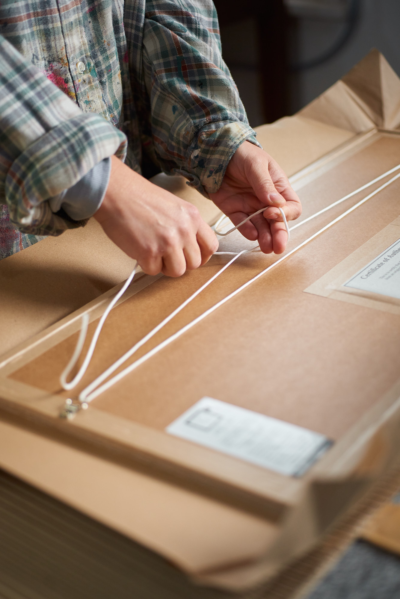 close-up image of hands tying nylon string on the reverse of a picture frame resting in brown paper packaging